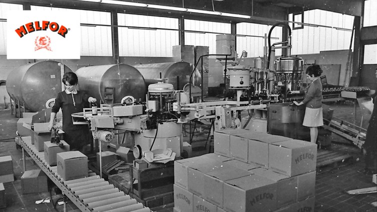 Produktionshalle mit Abfüll- und Verpackungsanlage, Kartons mit „Melfor“-Aufdruck und großen Tanks im Hintergrund.