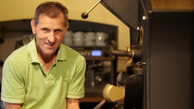 Person in grünem Polohemd steht neben einer schwarzen Kaffeeröstmaschine mit goldenen Details, im Hintergrund Tassen.