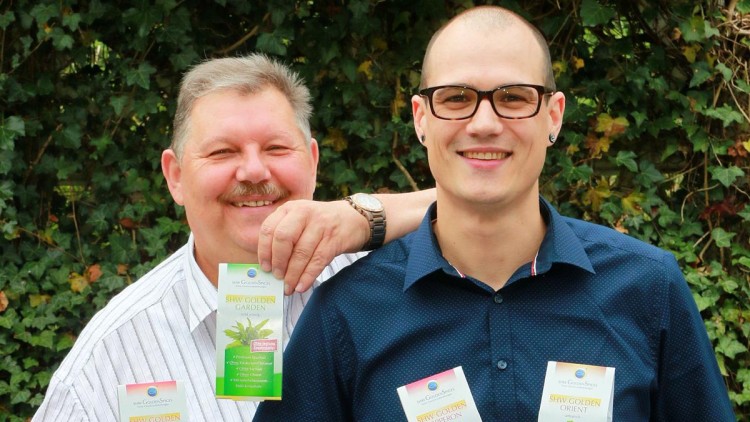 Zwei Personen halten und tragen grüne und bunte Verpackungen mit der Aufschrift „SHW GOLDEN GARDEN“ vor einer grünen Hecke.