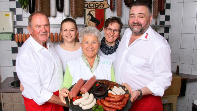 Eine Gruppe von Menschen in weißer Kleidung hält eine Platte mit verschiedenen Wurstsorten vor einem Hintergrund mit hängenden Würsten.
