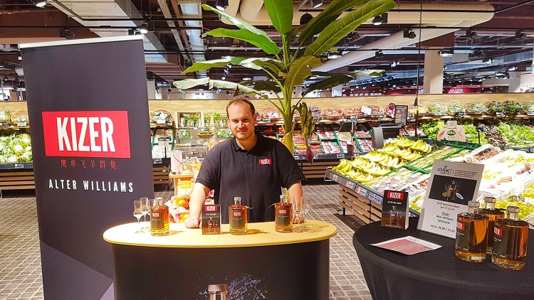 Ein Verkostungsstand mit KIZER-Produkten, Flaschen und Prospekten vor einer Obst- und Gemüseabteilung im GLOBUS-Markt.