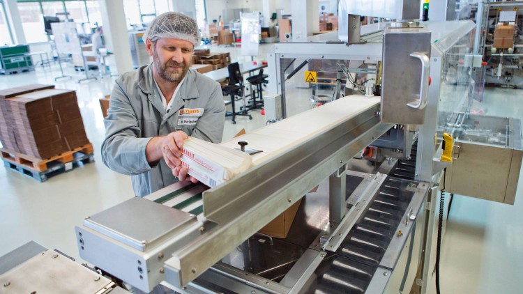Person in grauer Arbeitskleidung legt Packung auf Förderband einer Verpackungsmaschine, im Hintergrund Paletten und Kartons.