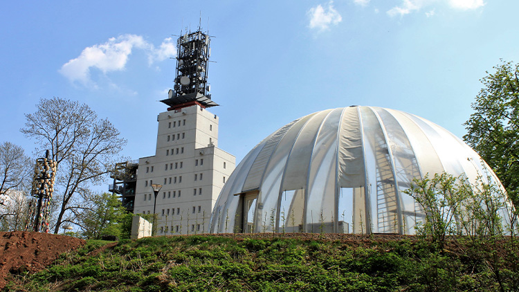 Eine weiße, kuppelförmige Zeltkonstruktion steht neben einem hohen Gebäude mit Antennen vor einem blauen Himmel und Bäumen.