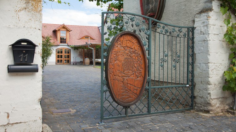 Ein geöffneter grüner Metallzaun mit Holzemblem „Weingut Bonnet“ führt zu einem Hof mit Haus und roten Dachziegeln.