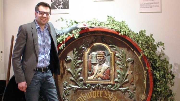 Großes hölzernes Bierfass mit geschnitztem Königsgesicht und Hopfendekoration vor einer weißen Wand mit Infotafeln.