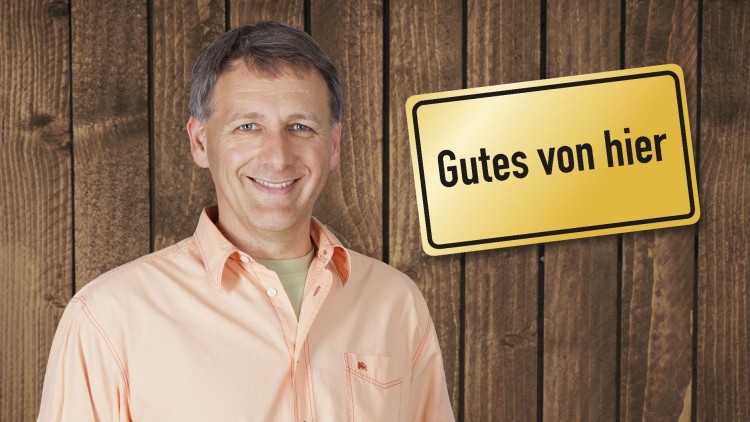 Person in orangefarbenem Hemd hält einen runden Käse vor Holzwand, daneben Korb mit Käseprodukten und Schild „Gutes von hier“.