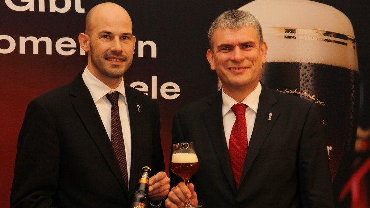 Zwei Männer in Anzügen halten eine Bierflasche und ein Glas mit Bier und weißem Schaum, im Hintergrund ein Schriftzug.