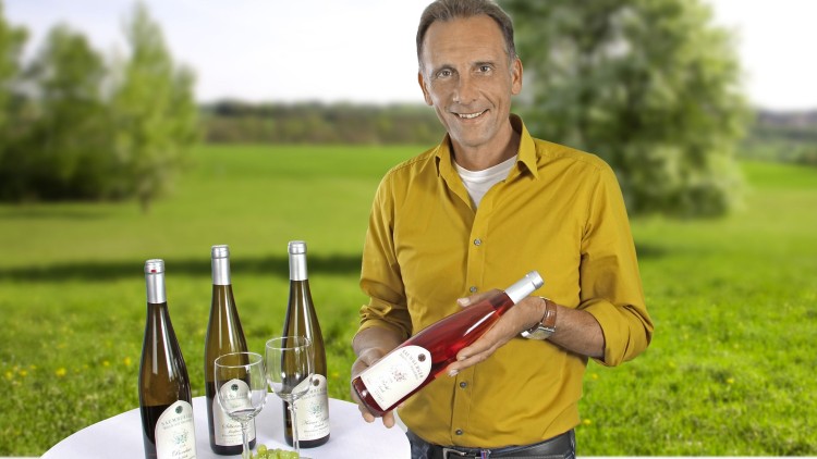 Mann in gelbem Hemd hält eine Flasche Roséwein, auf einem Tisch stehen Weißweinflaschen, Gläser und grüne Trauben.