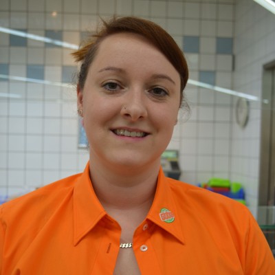 Person mit orangefarbener GLOBUS-Uniform, Namensschild „Susan Thomas“ und grünem GLOBUS-Pin vor heller Kachelwand.