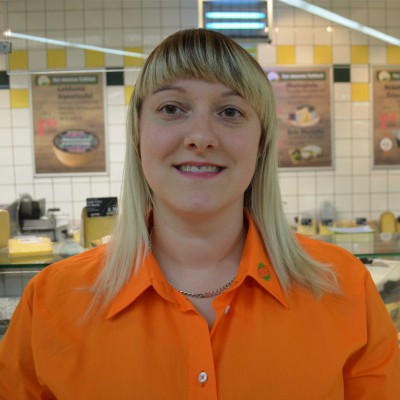 Person in orangefarbener Arbeitskleidung mit GLOBUS-Logo steht vor einer Käsetheke in einem Marktbereich mit Plakaten im Hintergrund.