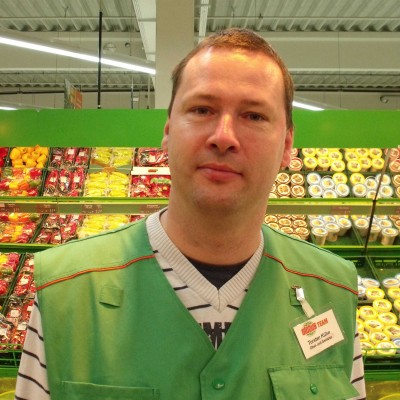 Person in grüner Arbeitsweste mit Namensschild steht vor Regalen mit Obst und gelb verpackten Lebensmitteln in einem Markt.