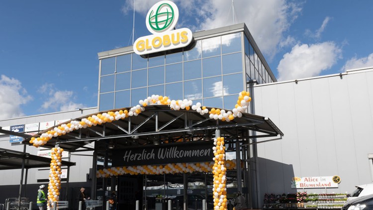 Eingang eines GLOBUS-Marktes mit Schriftzug „Herzlich Willkommen“, gelb-weißer Ballondekoration und Blumenverkauf rechts.