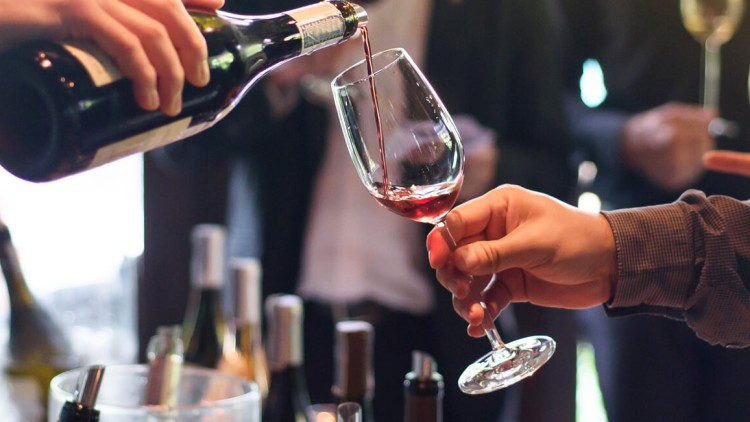 Eine Hand schenkt roten Wein aus einer Flasche in ein Glas, im Hintergrund stehen weitere Flaschen und Gläser.