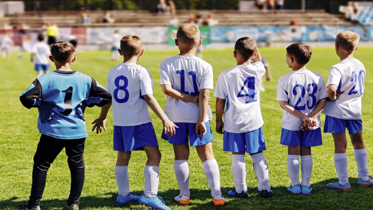 Sechs Kinder in weißen Trikots mit blauen Rückennummern stehen auf einem Fußballplatz und blicken auf das Spielfeld.