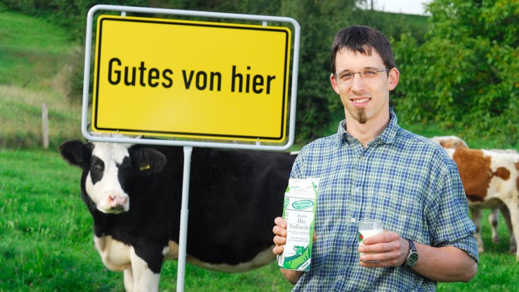 Ein Mann hält eine Packung Bio-Vollmilch und ein Glas Milch, im Hintergrund eine Kuh und ein gelbes Schild mit „Gutes von hier“.