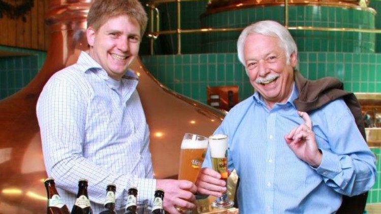 Zwei Personen stoßen mit Biergläsern an, umgeben von grünen Getränkekisten mit Großwald-Logo in einer Brauerei.