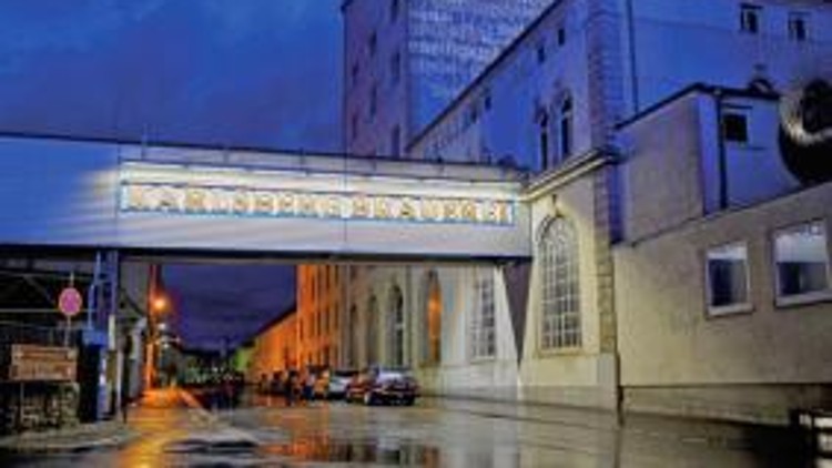 Ein beleuchtetes Industriegelände bei Nacht mit einer beschrifteten Brücke und einem nassen Straßenbelag im Vordergrund. 