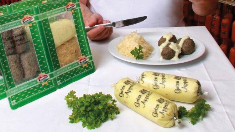 In einem Raum mit Würsten, Teller mit Knödeln und Sauerkraut, Packungen „Grüne Apfelknödel“ und Schild „Gutes von hier“.