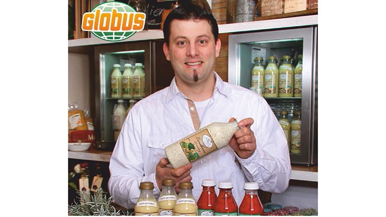 Person in weißem Hemd hält eine Packung Reis vor Regalen mit Getränken und Gewürzen, GLOBUS-Logo oben links sichtbar.