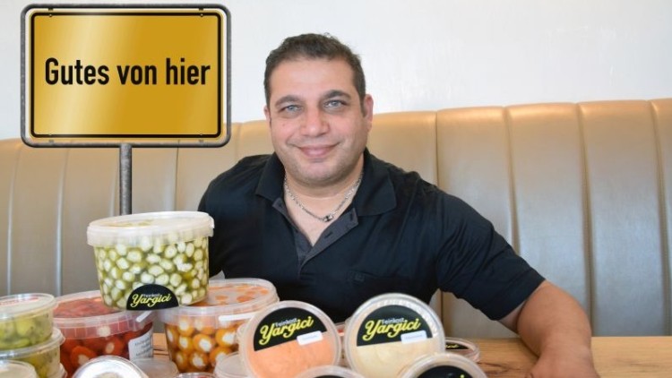 Eine Vielzahl an Plastikspeisenbehältern mit der Aufschrift „Yazgici“ steht auf einem Holztisch vor einer gepolsterten Sitzbank, Schild „Gutes von hier“.