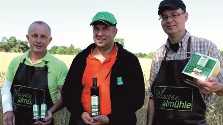 Drei Personen in schwarzen Schürzen halten Produkte mit grünen Etiketten der Marke „Bliesgau Ölmühle“ vor einem Feld.