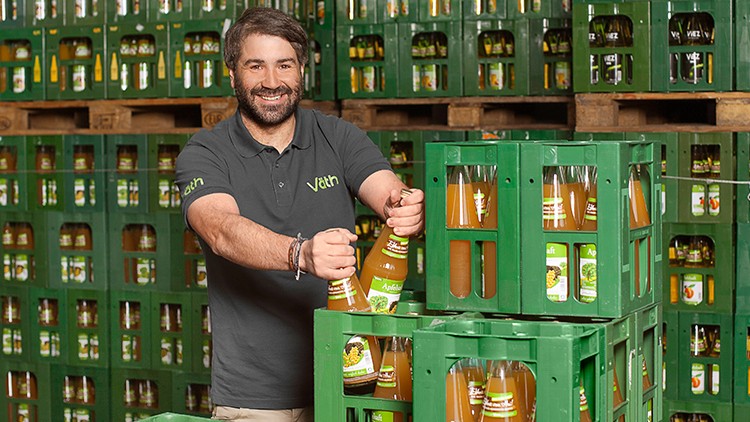 Person in grauem Hemd hebt eine Flasche Apfelsaft aus einer grünen Getränkekiste vor gestapelten Kisten.