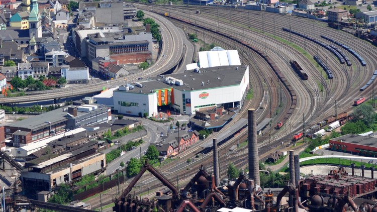 Ein GLOBUS-Markt mit weißer Fassade und bunten Streifen neben Bahngleisen, Industrieanlagen und Wohngebäuden.