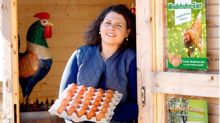 Person in blauer Weste hält eine offene Eierschachtel mit braunen Eiern vor einem Holzhaus mit Werbeschildern für Weidehuhn-Eier.
