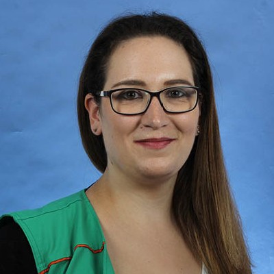 Person trägt eine grüne Arbeitsweste mit orangefarbenen Nähten vor einem hellblauen Hintergrund.