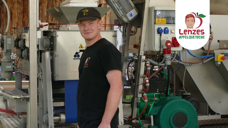 Industrielle Maschine mit Rohren, Motoren und Kabeln vor Holzwand, Text auf T-Shirt: Lenzes Apfelquetsche.