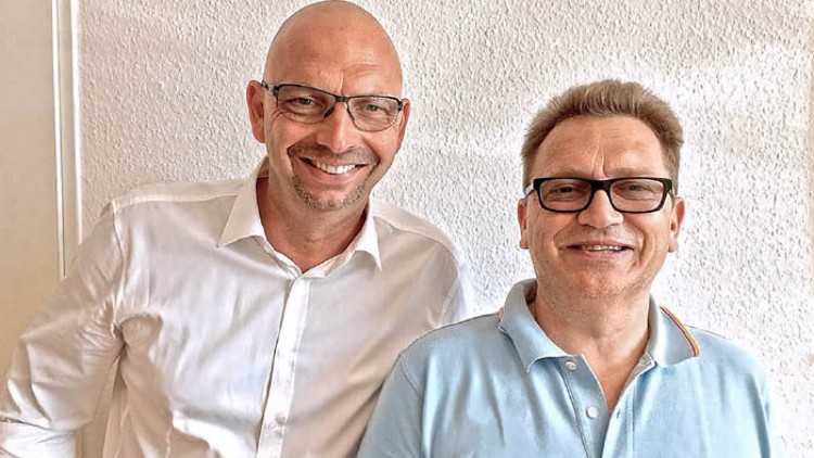 Zwei Personen in Hemden stehen vor einer hellen Wand, eine Person trägt ein weißes Hemd, die andere ein hellblaues Polohemd.