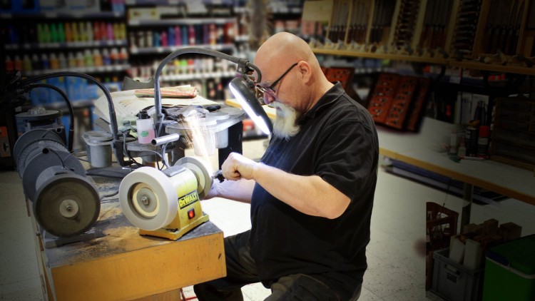 Person arbeitet mit einem Schleifgerät an einer Werkbank, umgeben von Werkzeugen und Regalen mit bunten Produkten.
