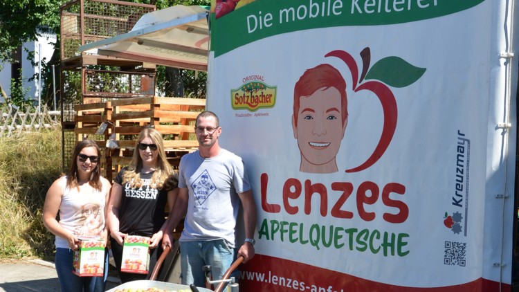 Drei Personen stehen vor einer mobilen Kelterei mit der Aufschrift „Lenzes Apfelquetsche“, davor eine Wanne mit Äpfeln.