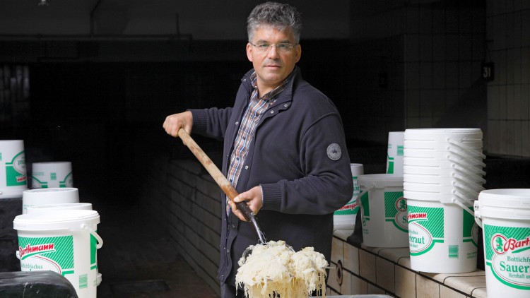 Person rührt mit einer Holzstange in einer Wanne mit Sauerkraut, daneben stehen weiße Eimer mit grünem Aufdruck.