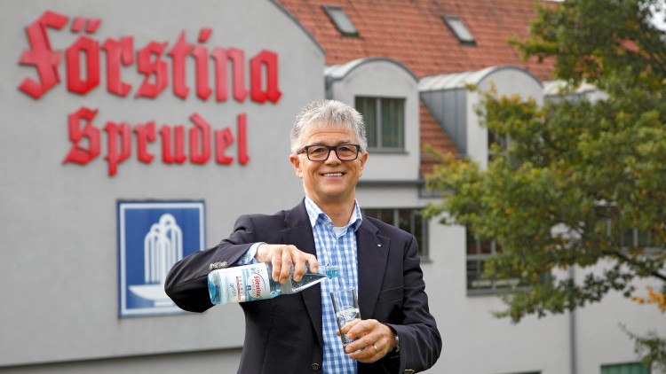 Eine Person in Sakko und kariertem Hemd gießt Förstina Sprudel aus einer Glasflasche in ein Trinkglas, Hintergrund mit Firmengebäude.