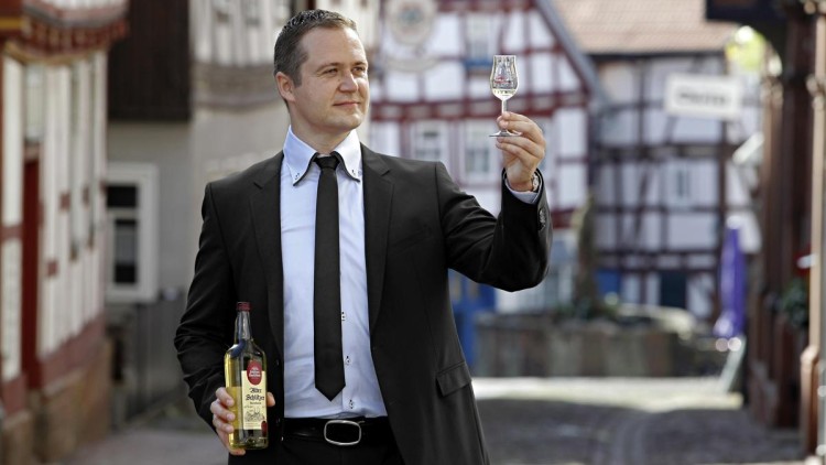 Person im Anzug hält eine Flasche „Alter Schlitzer“ und ein gefülltes Glas in einer Altstadt mit Fachwerkhäusern.