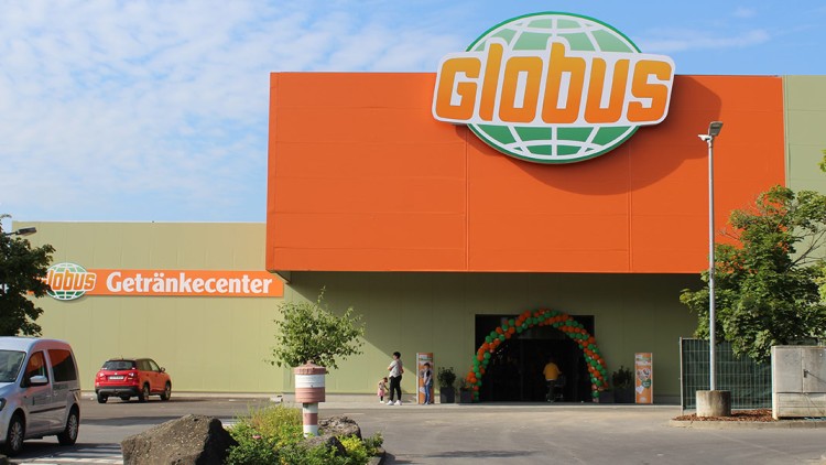 Eingangsbereich eines GLOBUS Getränkecenters mit orangen und grünen Ballons, Autos, Zebrastreifen und einer bepflanzten Verkehrsinsel.