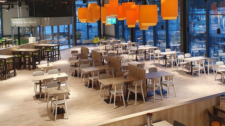 Ein moderner, heller Gastronomiebereich mit Holztischen, Stühlen, orangen Lampen und einem Notausgangsschild an der Decke.