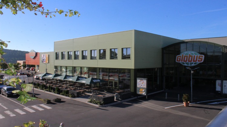 Das GLOBUS-Einkaufszentrum mit grünem Gebäude, Glasfassade, Terasse mit Sonnenschirmen und Parkplatz bei klarem Himmel.