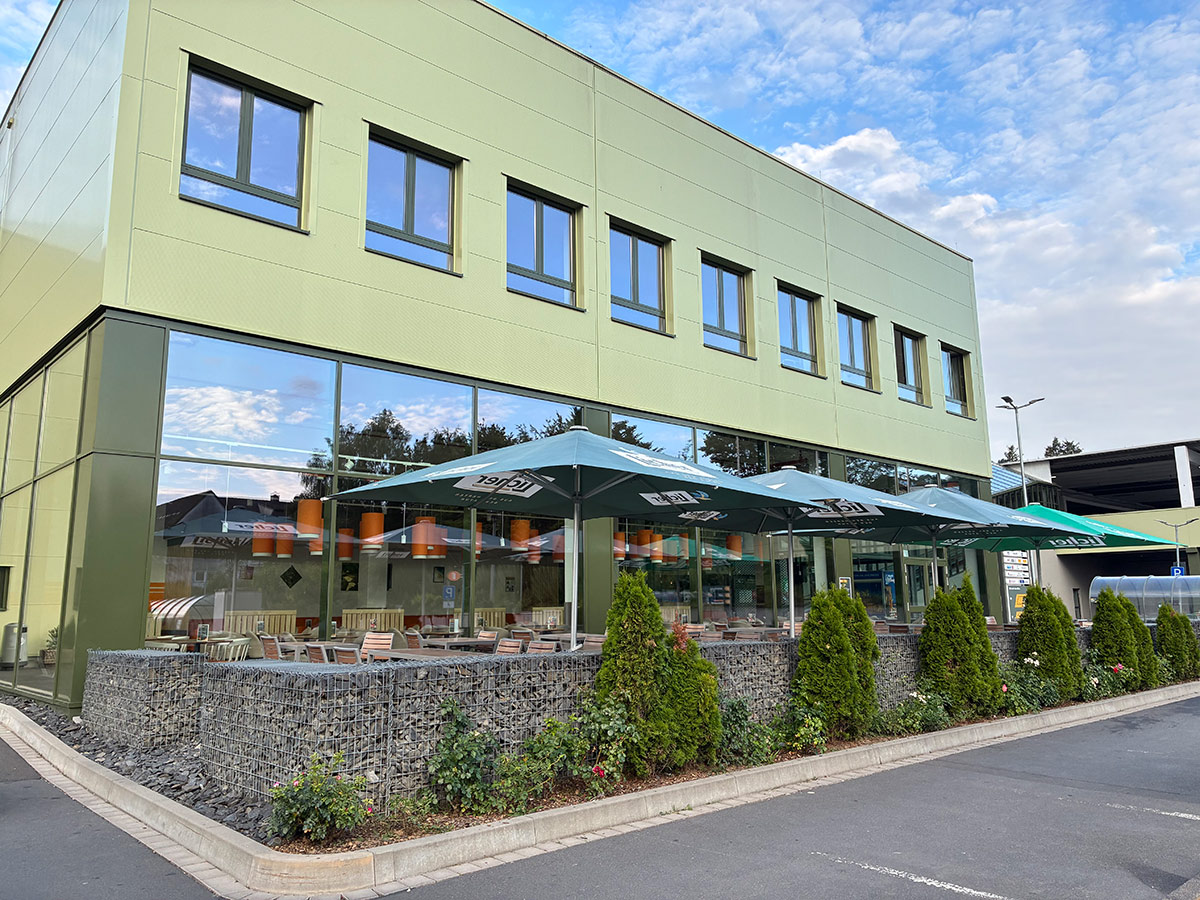 Ein grün-graues Gebäude mit Glasfront, davor Sonnenschirme mit GLOBUS-Logo, Sitzbereich und Pflanzen in Steinbegrenzung.