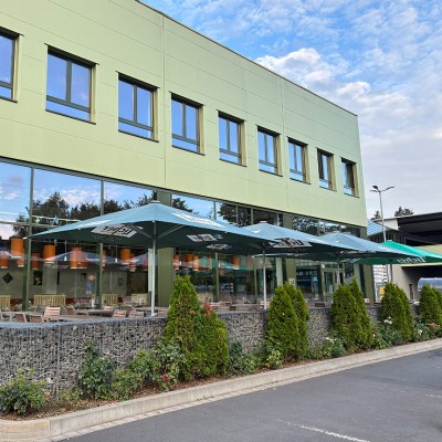 Ein grün-graues Gebäude mit Glasfront, davor Sonnenschirme mit GLOBUS-Logo, Sitzbereich und Pflanzen in Steinbegrenzung.