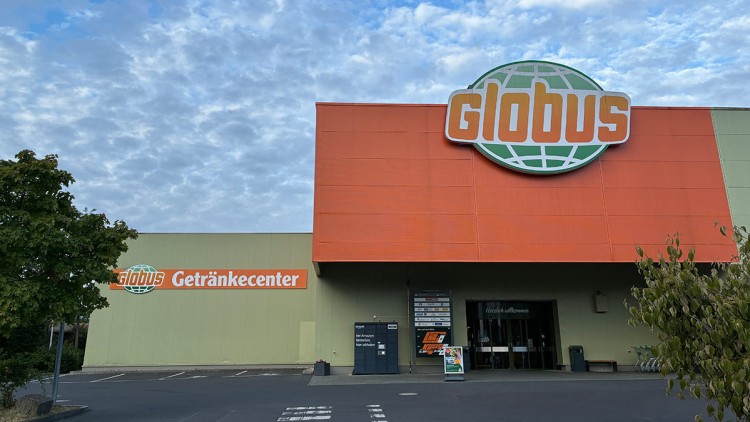 Eingangsbereich eines GLOBUS Getränkecenters mit auffälliger orangegrüner Fassade unter blauem, wolkigem Himmel.