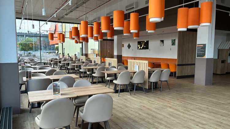 Großer, heller Speiseraum mit Holzfußboden, grauen Stühlen, Holztischen, orangefarbenen Lampenschirmen und moderner Einrichtung.