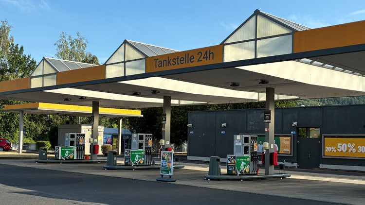 Eine überdachte Tankstelle mit der Beschriftung „Tankstelle 24h“, Zapfsäulen und Werbeschildern mit Rabattangaben vor einem Gebäude.