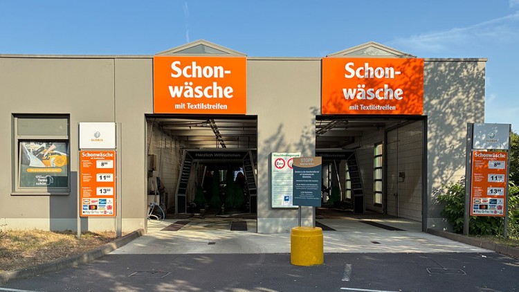 Zwei Waschstraßen mit orangefarbenem „Schonwäsche mit Textilstreifen“-Schriftzug, Schilder mit Infos und Preisen davor.