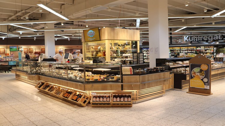 Käsetheke im GLOBUS-Markt mit Auswahl an Käsesorten, Regalen mit Weinflaschen und Aufsteller mit Werbetext.