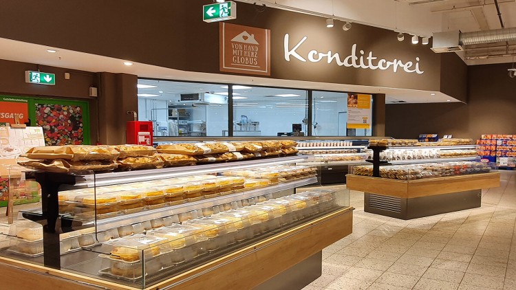 Eine helle GLOBUS-Konditorei mit Glasvitrinen voller Kuchen, Torten und Gebäck, darüber das Schild „Von Hand mit Herz GLOBUS“.