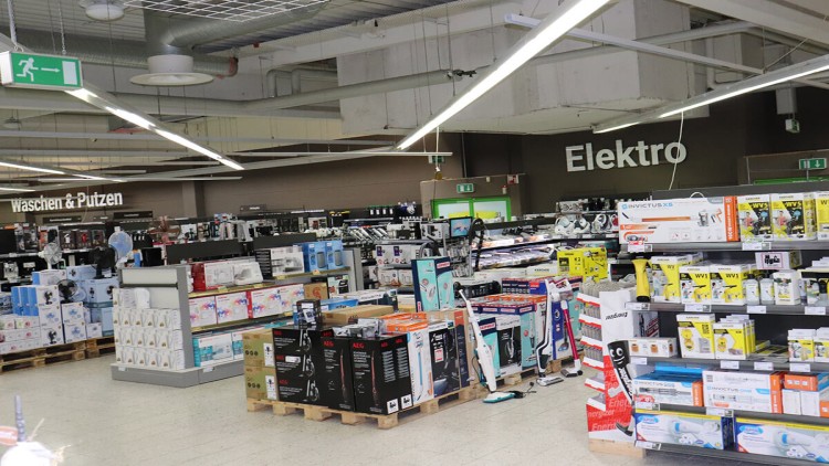 Regale und Paletten im GLOBUS-Markt, gefüllt mit Elektroartikeln und Reinigungsgeräten, Beschilderung „Waschen & Putzen“ und „Elektro“.