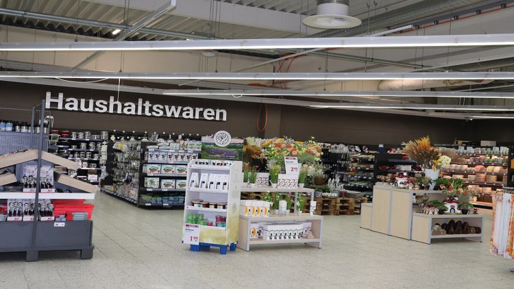Regale und Verkaufsflächen mit Haushaltswaren, Dekorationen und Blumenarrangements, darüber ein braunes Schild mit „Haushaltswaren“.