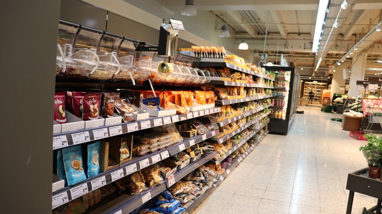 Regale mit verpackten Lebensmitteln wie Snacks und Trockenfrüchten, Preisschilder sichtbar, helle Supermarktbeleuchtung.